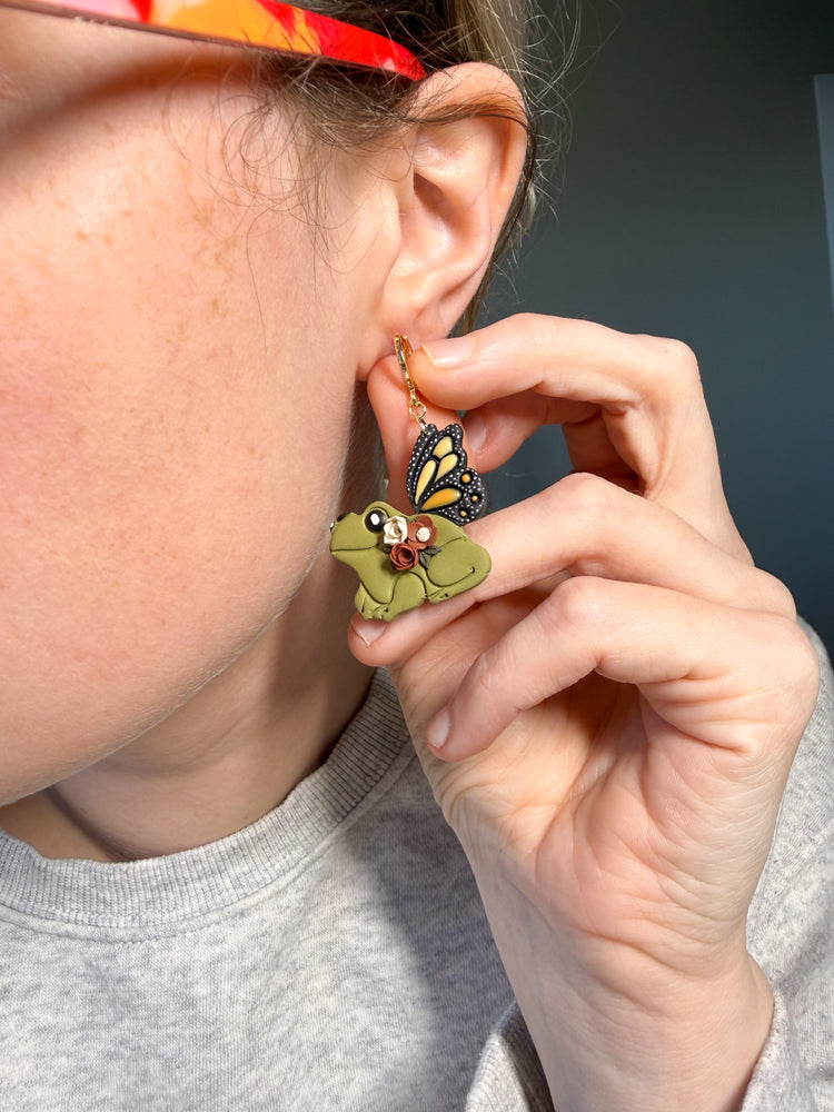 Person wearing a unique earring with a green frog and butterfly design, holding it near their ear.