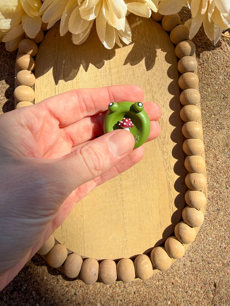 Hand holding a small green frog figurine in front of a wooden frame with flowers.