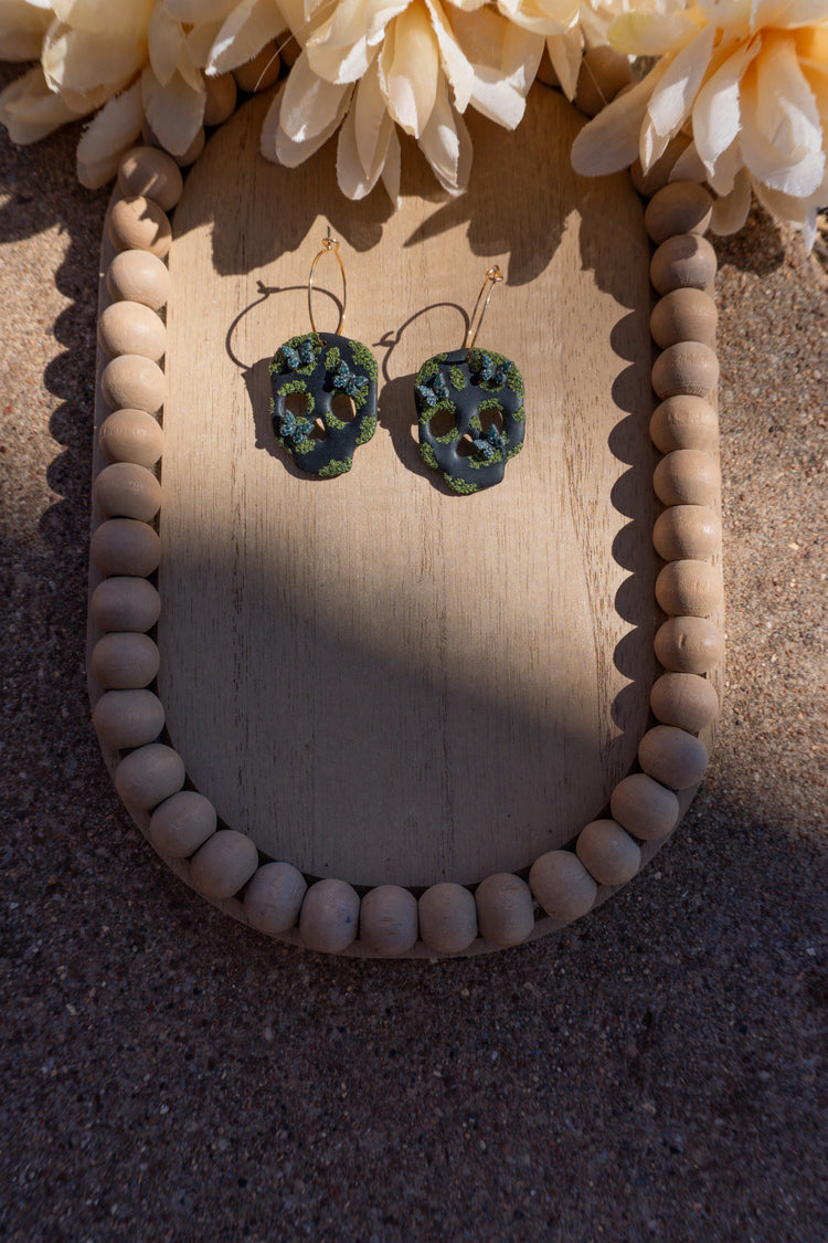 Skull earrings that are cottage-core themed, attached to gold hoop earring posts.