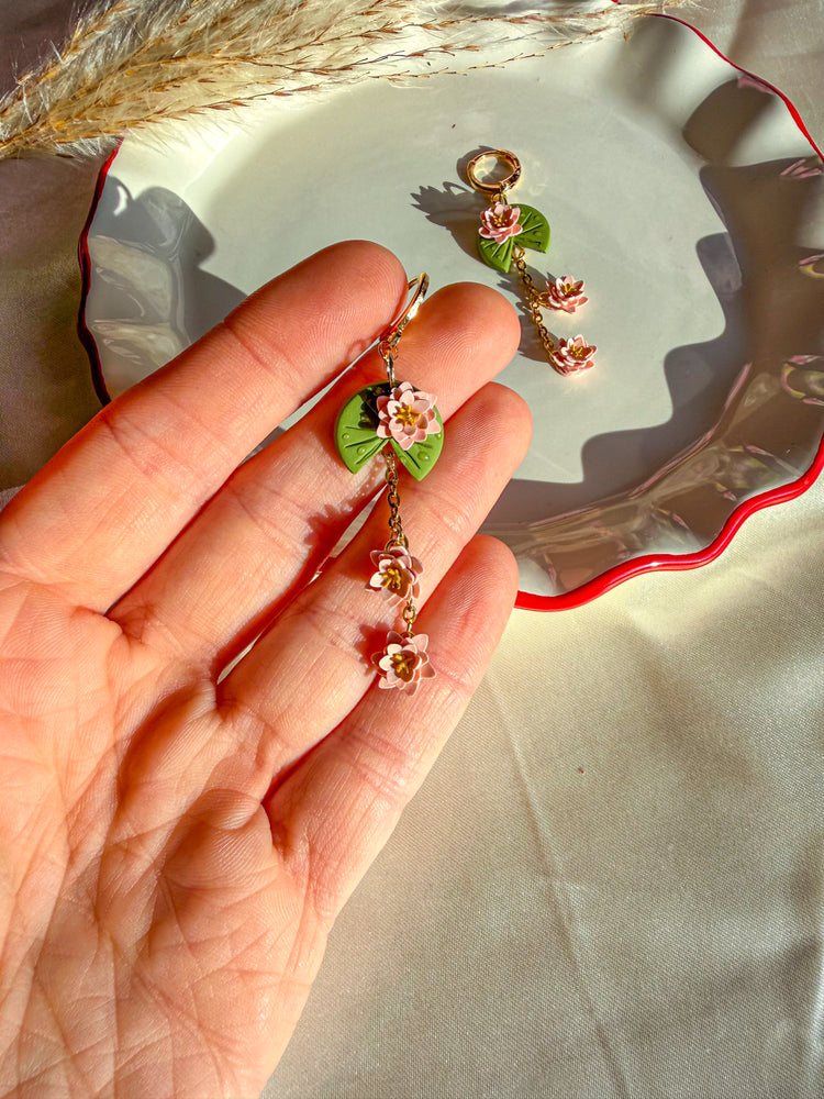 Hand holding a pair of lily pad floral earrings with a decorative plate in the background