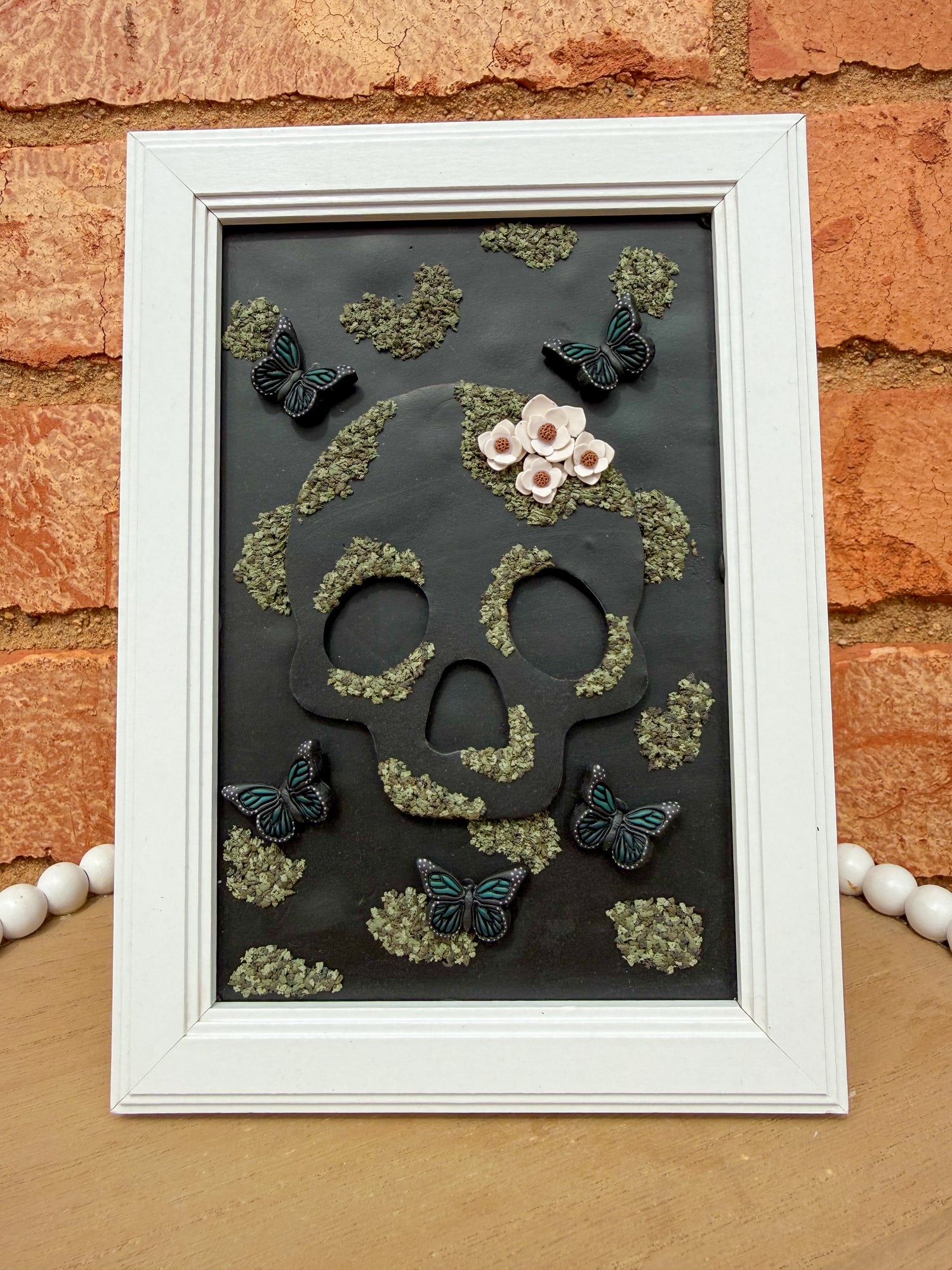 Decorative skull and floral design in a white frame against a brick wall.
