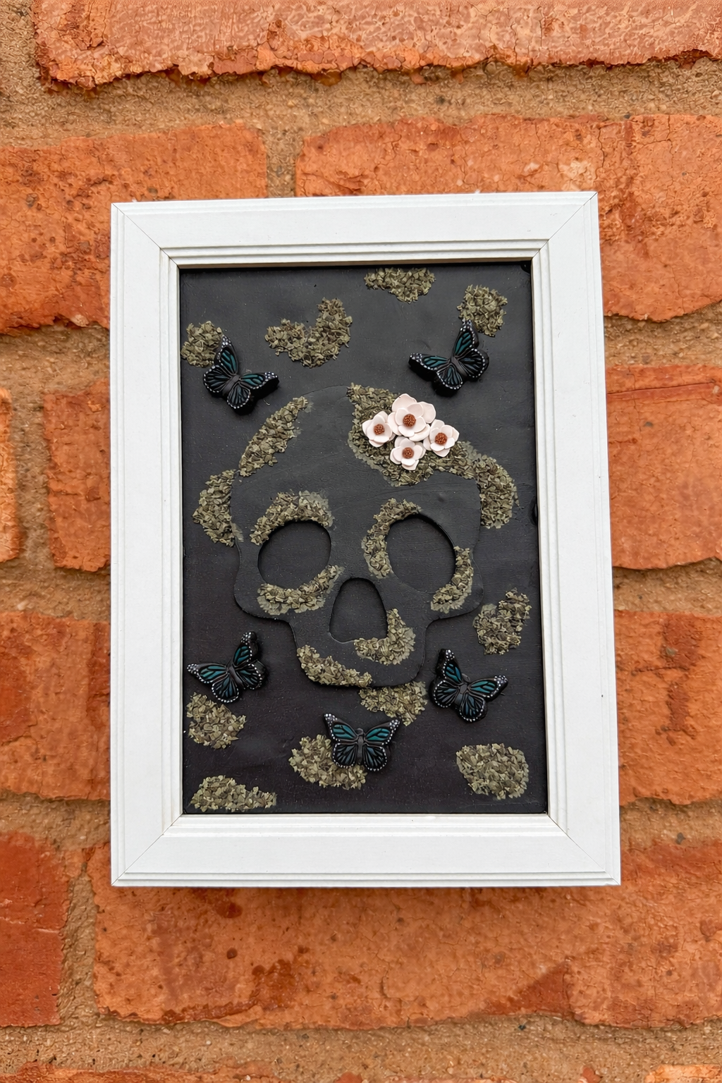 Handmade decorative wall art with skull design, moss, and butterflies hanging on a brick wall