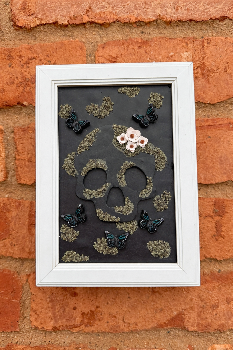 Handmade decorative wall art with skull design, moss, and butterflies hanging on a brick wall