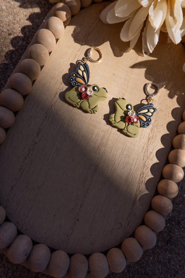 Frog wearing butterfly wings and flowers dangle earrings with gold lever-back earring posts.
