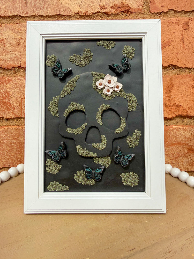 Polymer clay framed artwork with a skull design, moss, flowers, and butterflies on a brick wall background.