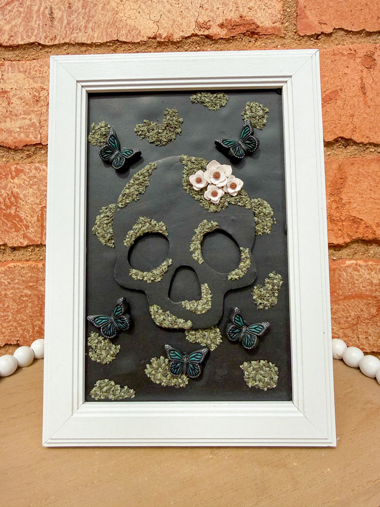 White frame skull polymer clay art piece leaning against a brick wall.