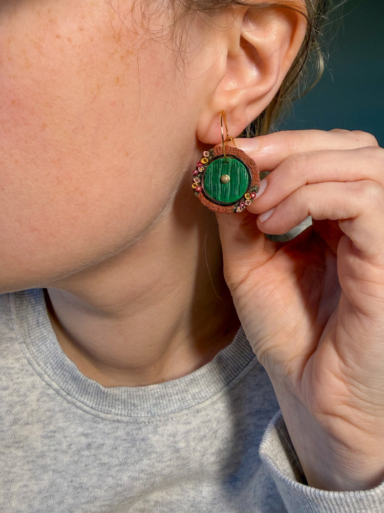Close-up of a person wearing a green and brown  hobbit door earring. On the hobbit door there are colorful wildflowers. 